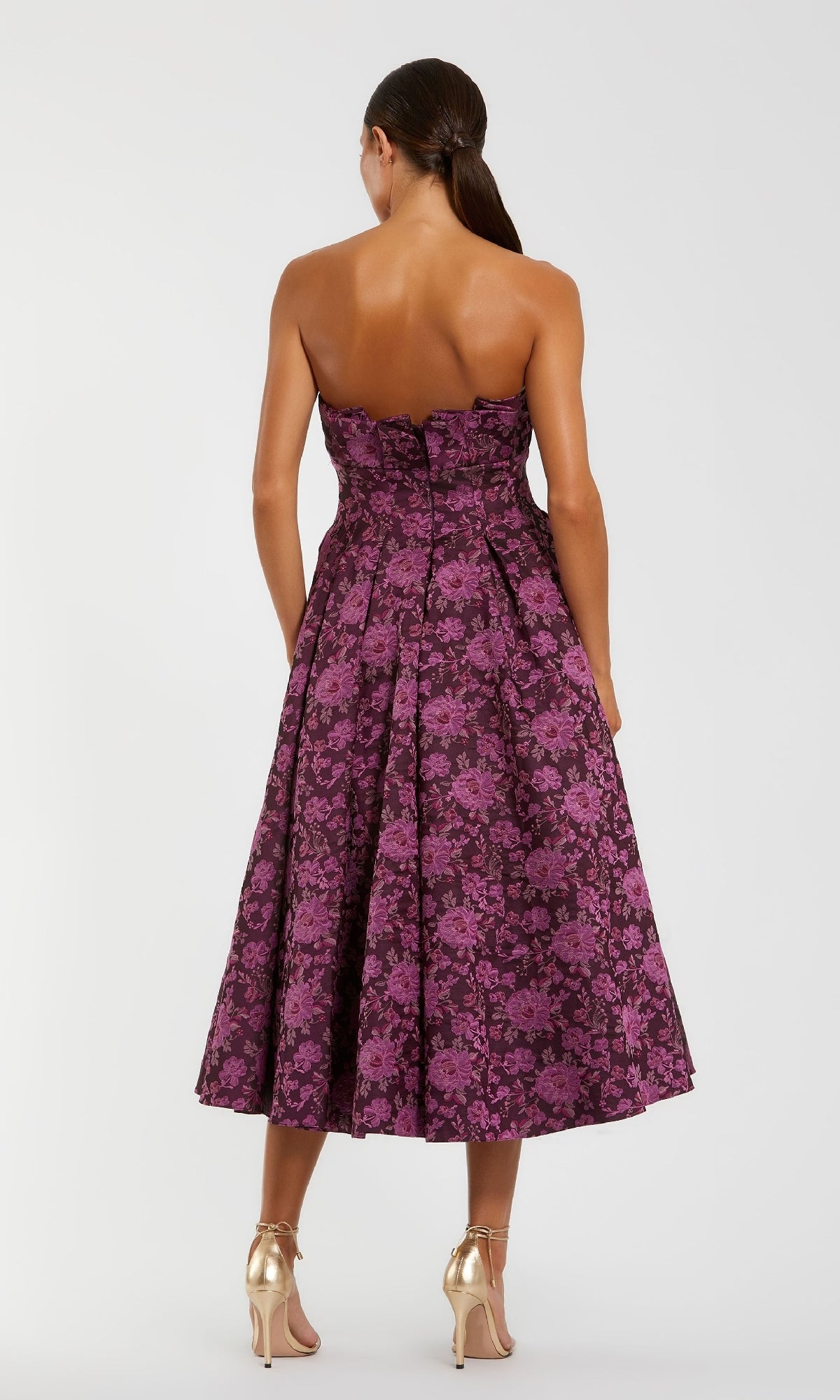 A woman faces away in the Mac Duggal 56159 Strapless Wedding Guest Dress, a floral-print midi paired with gold heels—an elegant choice for wedding guests.